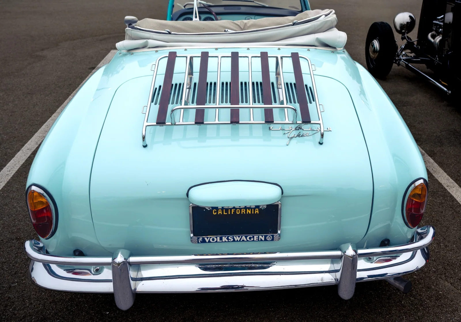 Rear of the seafoam Karmann Ghia convertible, showing its folded light-colored roof, chrome luggage rack with dark slats, Karmann Ghia script, and vertical tail lamps.